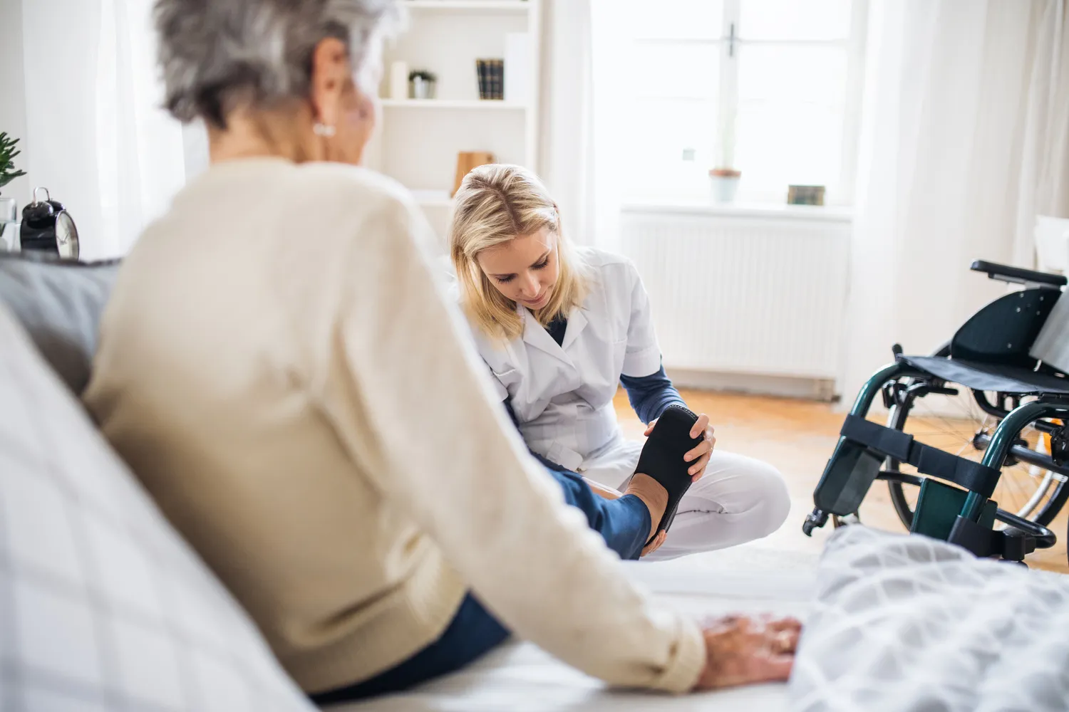 Care Comfor GmbH - Pflegedienst - Ein Gesundheitsbesucher zieht einer Seniorin zu Hause Hausschuhe an.